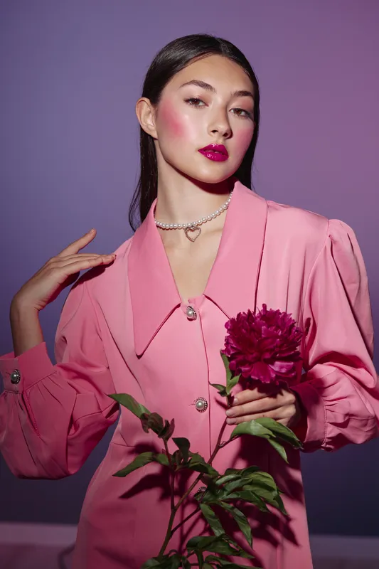 Young woman in a pink blouse poses against a purple background holding dark pink peony. Bold fuchsia lipstick, soft lighting, creative beauty fashion concept for modern style.