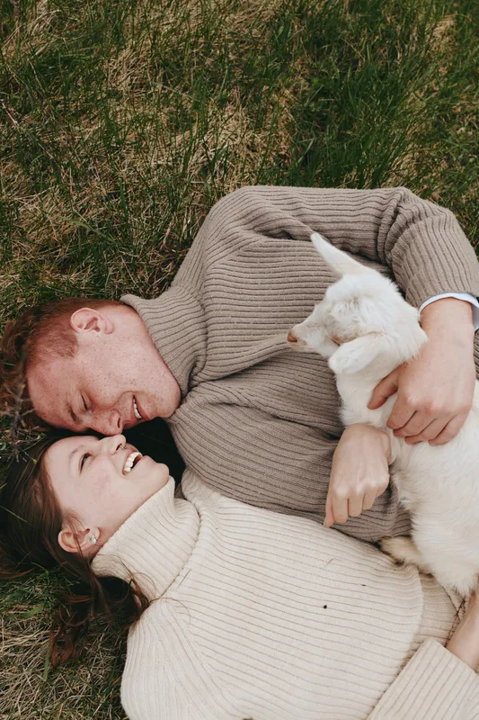 A joyful couple lies on grass, cuddling a goat kid, sharing a tender outdoor moment, surrounded by warm sunlight and soft sweaters, capturing simple, everyday happiness and connection.