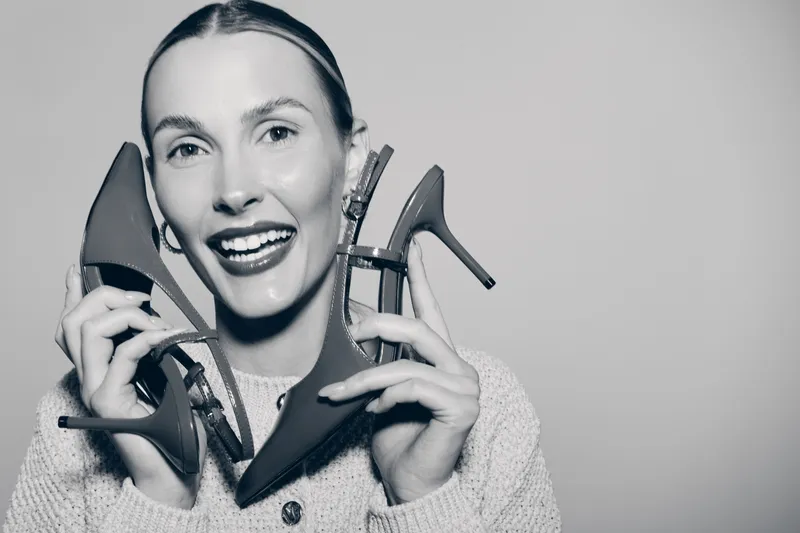 Smiling caucasian woman with fair skin holding high heel shoes near face. Fashion concept with happy female model and stylish footwear isolated on plain background.