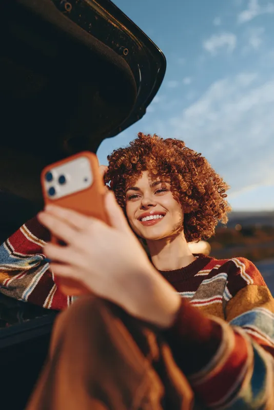 Woman lifestyle selfie smartphone curly hair smiling outdoors car window, young woman grins while holding a smartphone close to the car seat, warm sunlight, travel mood