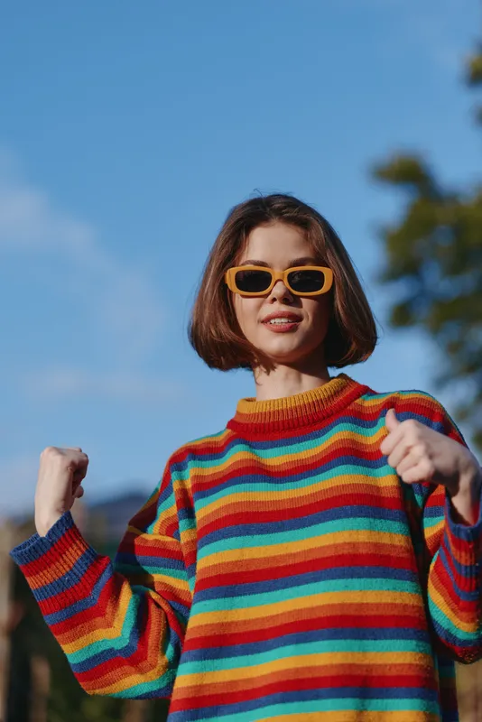 Woman in rainbow sweater and yellow sunglasses smiling outdoors in a confident portrait pose with raised fists, short bob haircut, vibrant colorful fashion vibe and sunny blue sky backdrop.