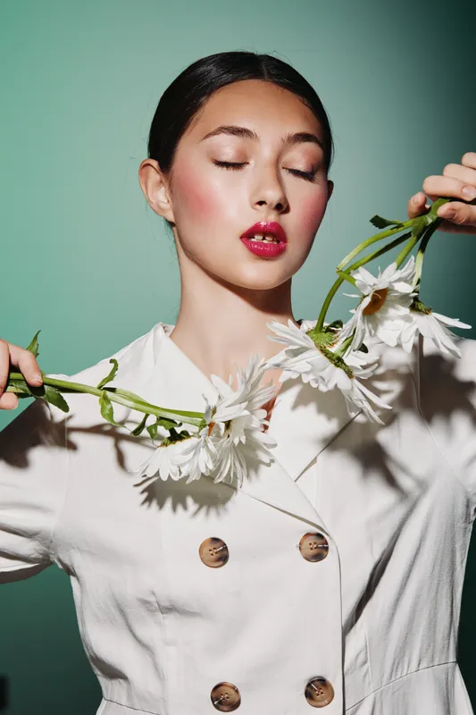 Young woman with sleek hair and red lipstick holds white flowers posing creatively in a double-breasted cream jacket. Editorial studio shot with soft textured background.