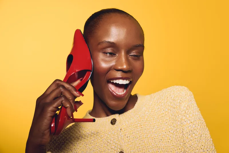 Happy African woman with dark skin laughs holding red high heel shoe near ear as if talking on phone. Yellow background, casual outfit. Concept of joy, fashion, and playful communication.