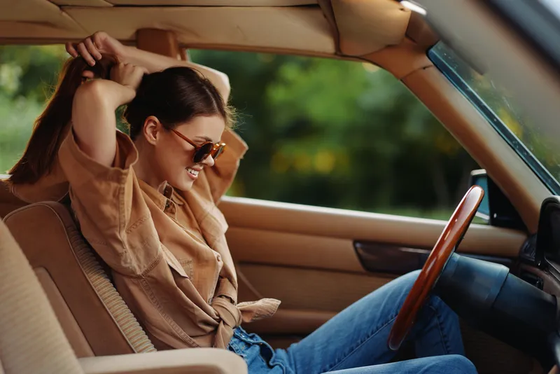 Young woman smiling while adjusting her hair in a vintage car, wearing stylish sunglasses and a casual outfit against a vibrant green background.