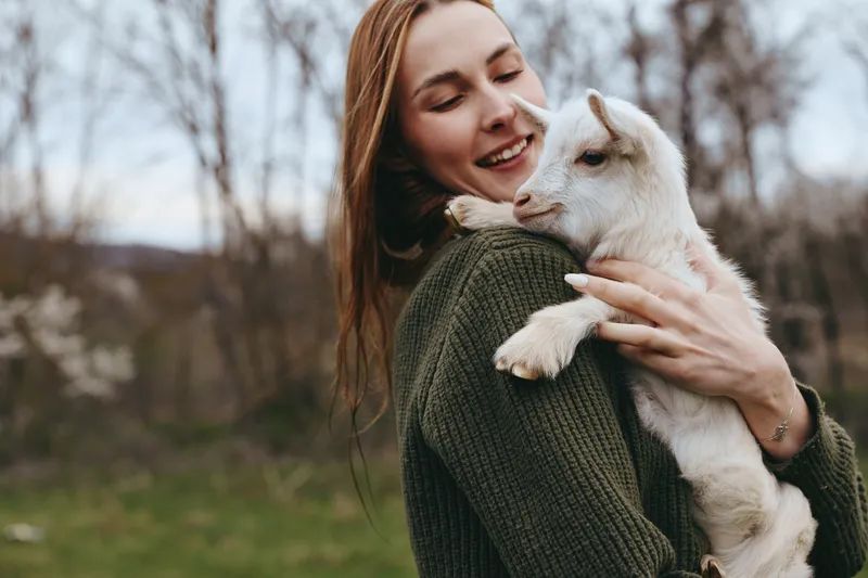 Young woman with long hair smiles gently while holding a small white baby goat outdoors in a natural setting during a calm autumn day.