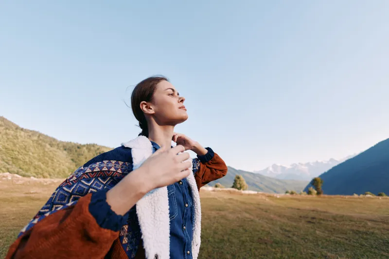 Woman standing in meadow with mountains behind, portrait of a traveler outdoors. Peaceful expression, sunlight on jacket and sweater, scenic landscape for calm relaxation.
