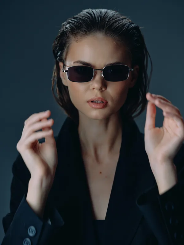Young woman wearing sunglasses and black blazer posing confidently in low light with modern style and serious expression