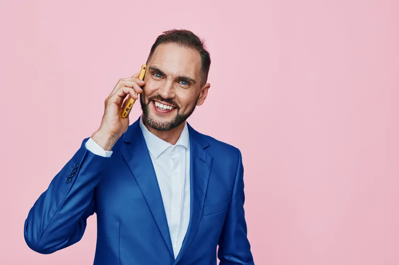 Professional man in a blue suit speaks on the phone against a solid pink background, projecting confident energy and friendly emotion in a clean studio setting with an isolated color backdrop.