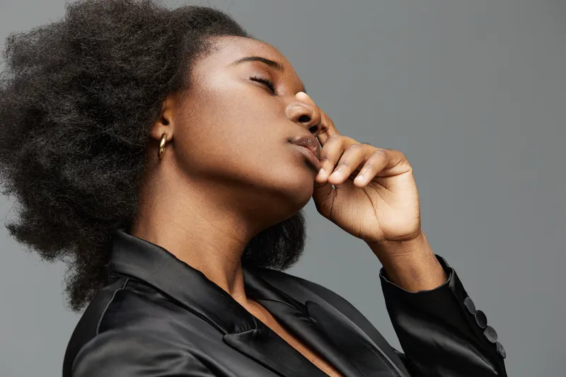 A thoughtful Black woman in a sleek black suit poses against a gray background, showcasing elegance and introspection, embodying confidence and serenity