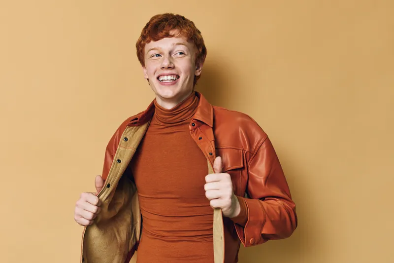 Young man with red hair wearing jacket and turtleneck smiling and showing thumbs up posing against beige background with cheerful and confident expression.
