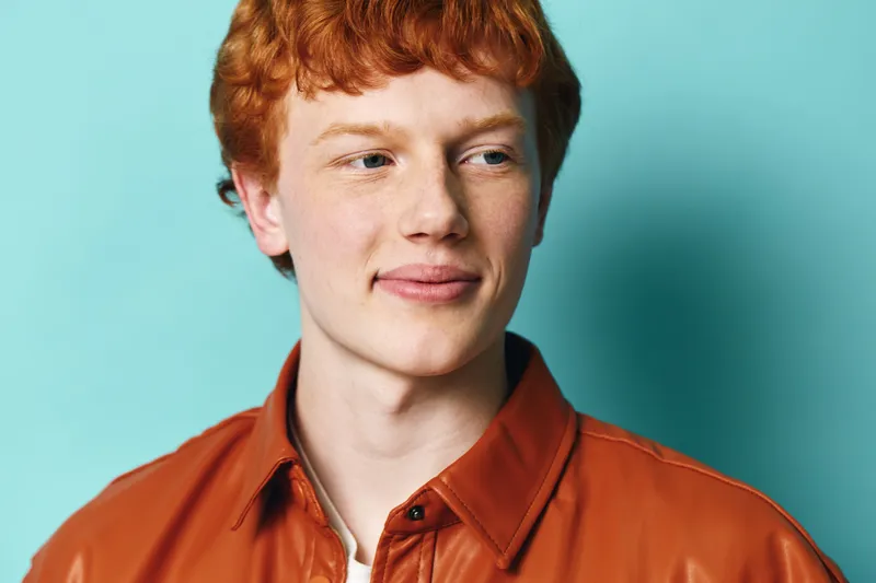 Young man with red hair and clear skin wearing brown leather jacket posing on blue background. Casual style portrait with natural light, confident look and relaxed mood in studio.