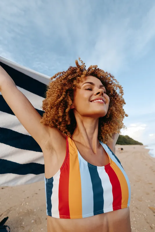 Smiling woman with curly hair on a sunny beach wearing a colorful striped swimsuit, arms raised, towel visible in the background. A vibrant scene of summer fun, vitality, and freedom.
