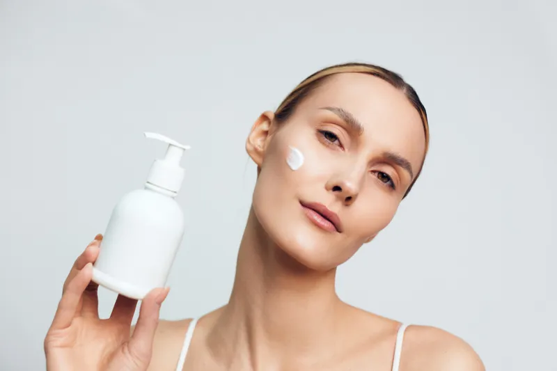 A young woman with clear skin applies lotion from a white pump bottle, posing in a bright studio to convey a calm skincare routine and self care moment.