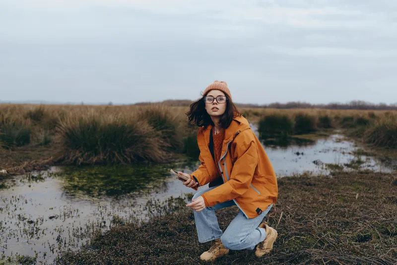Woman outdoor lifestyle kneeling on grassy riverbank wearing orange jacket in cloudy autumn landscape, natural scenery with soft daylight, calm thoughtful mood, nature and fashion