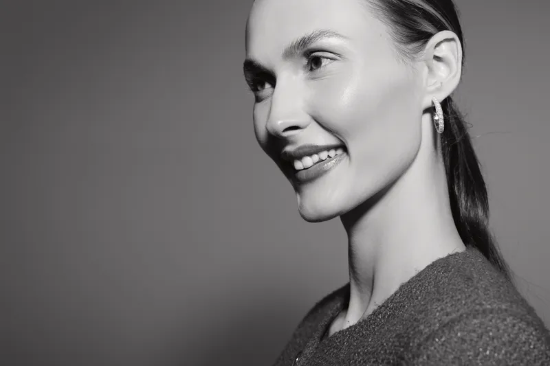 Smiling caucasian woman with light skin and dark hair looks away, wearing earrings and a sweater. Studio portrait with soft lighting and neutral background.