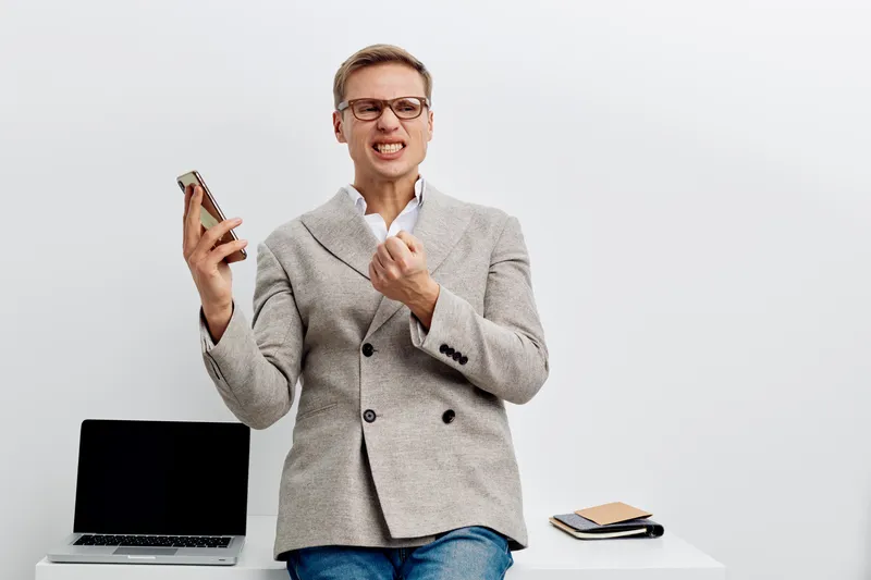 Confident young woman with short blonde hair, glasses, and grey blazer expressing frustration while holding smartphone, seated at modern office desk with laptop and notebooks, on plain white backgroun