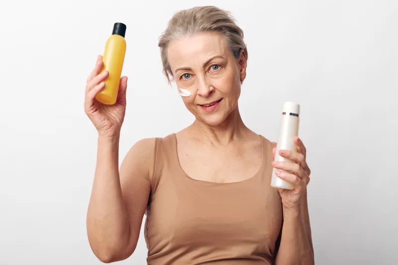 Mature woman with light skin and short hair applies cream on face, holding two skincare bottles against white background. Concept for beauty, wellness, and anti aging care.