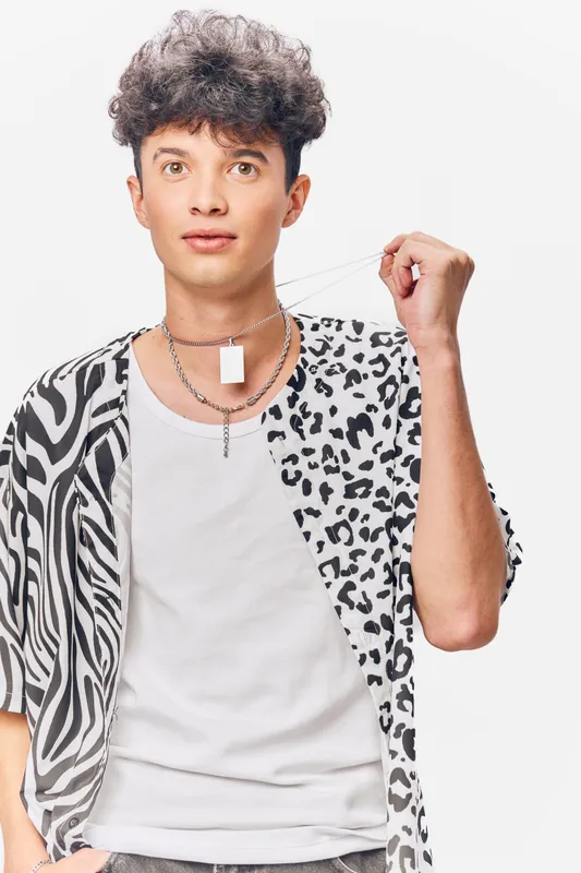 Young man wearing a patterned shirt and white tank top, holding a necklace pendant confidently against a plain white background, showcasing modern fashion style.