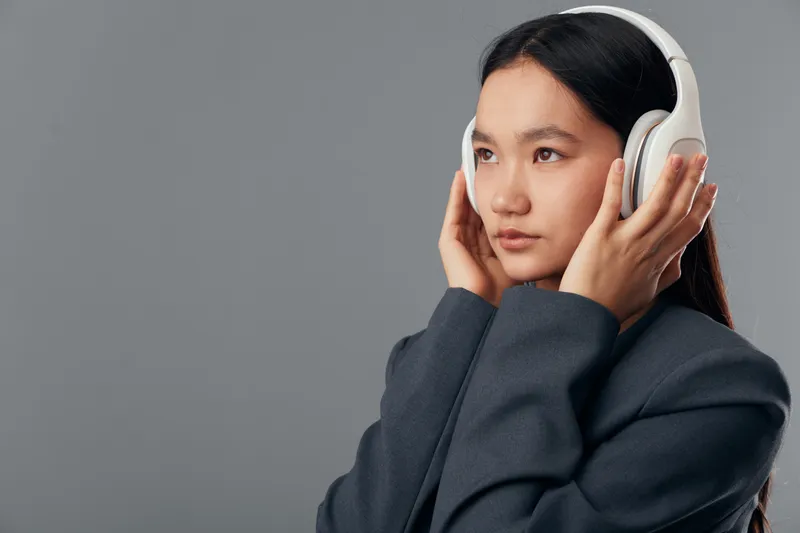 Young woman wearing large white headphones listening to music with focused expression against gray background in studio. Technology, audio, sound and lifestyle concept portrait.