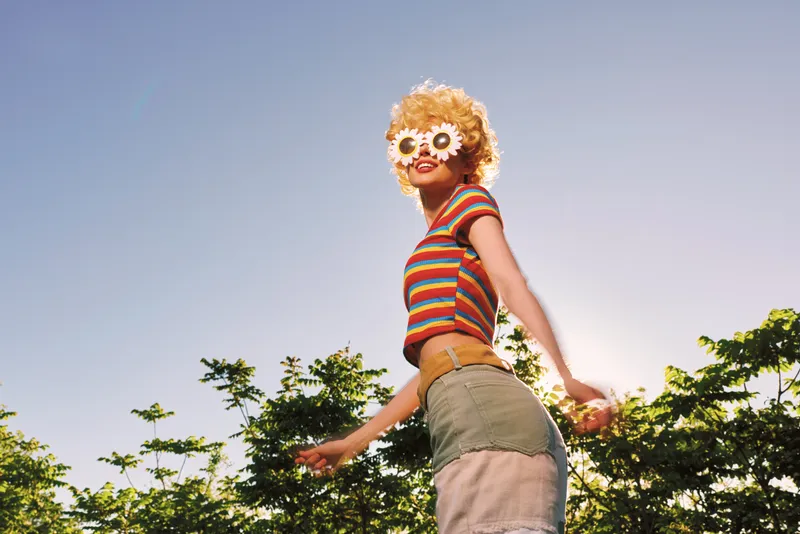 A woman wearing a striped top and shorts stands outdoors with flower glasses, embodying a carefree summer lifestyle with a retro vintage vibe, bright colors and sunny mood.