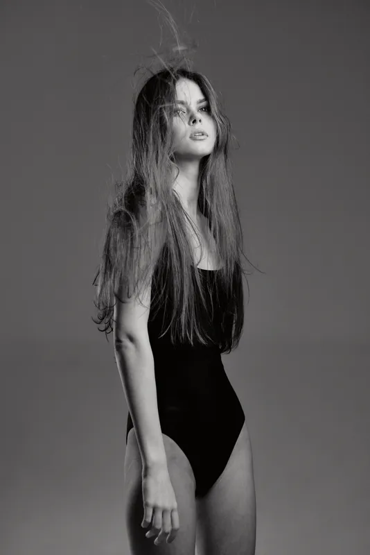 Black and white portrait of a young woman with long hair wearing a black swimsuit, expressing calm and confidence in studio lighting.