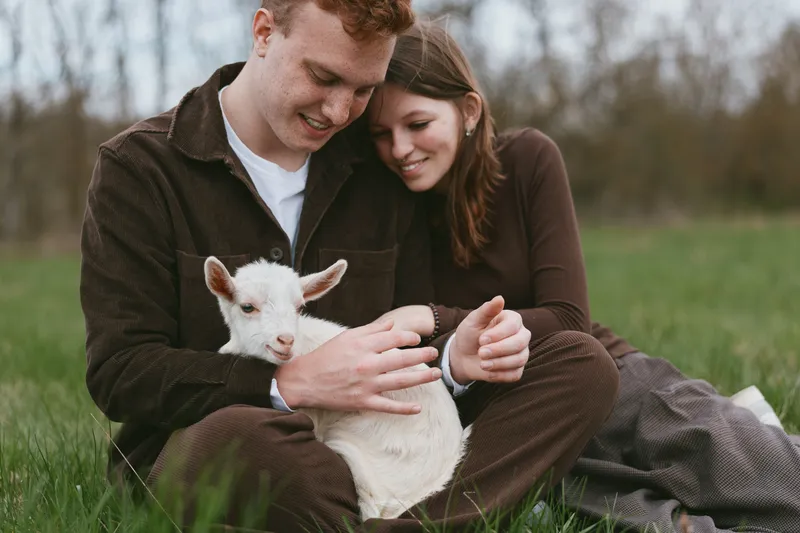 A young couple sits on grass holding a white baby goat while enjoying a quiet moment outdoors in casual brown clothing during an early spring day in nature.