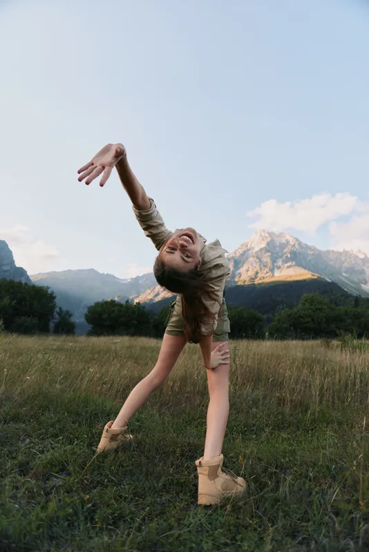 Young woman in beige outfit stretches and smiles joyfully in a mountain meadow during golden hour with clear sky and distant peaks in the background.