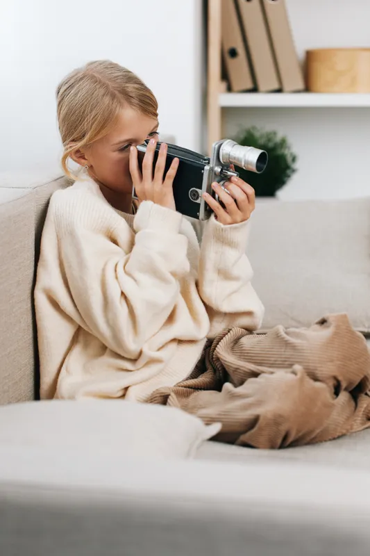 A young girl sits on a sofa wearing a soft sweater, holding a vintage camera to her eye as she discovers photography in a cozy, candid moment.