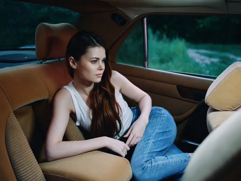 Thoughtful young woman in casual outfit sitting in car, reflecting on life in a serene natural setting, surrounded by greenery and soft colors.