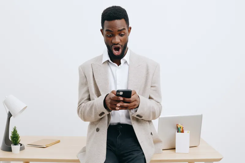 Surprised young African American man in beige blazer looking at mobile phone with shocked expressions while standing in modern office setting, desks, laptop, pens, and plant on white background. Peopl