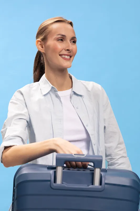 Smiling woman in casual white shirt with ponytail holds blue suitcase handle, looking away on blue background. Concept of travel, vacation, and holiday preparation.