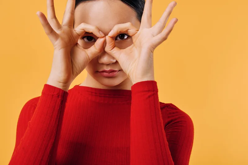Woman in red sweater with dark hair makes binocular gesture with hands around eyes against orange background. Closeup portrait with playful expression and creative pose for fun concept.