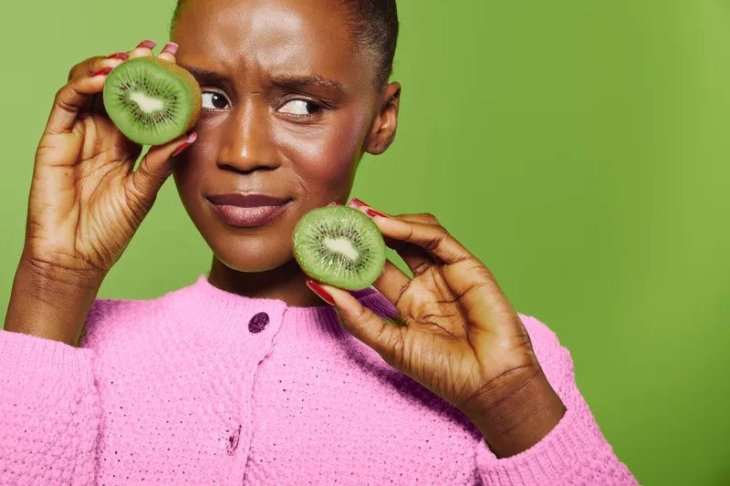 Young African woman with dark skin holds two halves of fresh kiwi near her face. She wears a pink sweater and poses on a bright green background. Concept of healthy food and natural beauty.