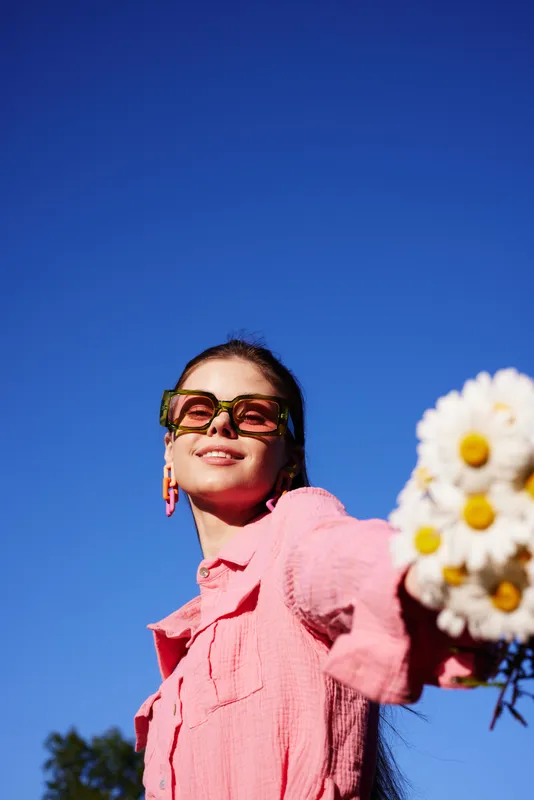 Young woman smiling with a bouquet of daisies, wearing stylish green sunglasses and a pink shirt against a bright blue sky Spring vibes, cheerful and vibrant