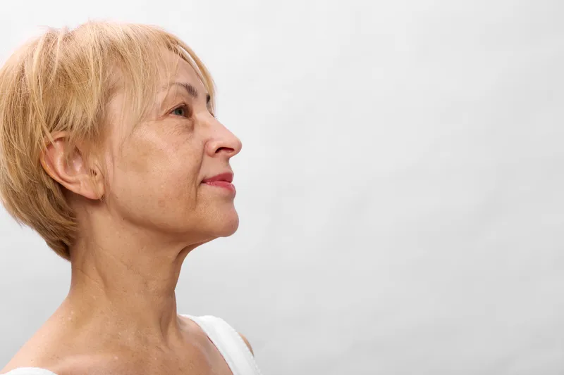 Senior woman profile with blond short hair, looking to the side, thoughtful expression, closeup face portrait, soft studio light, neutral background, calm mood, gentle skin texture