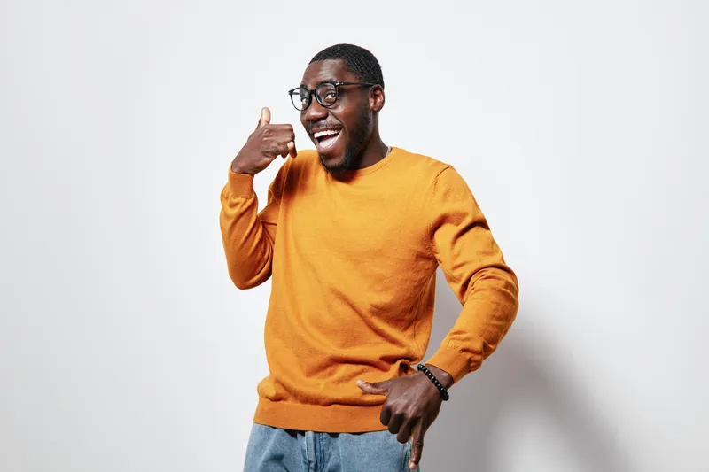 African American man with dark skin wearing glasses and orange sweater making call me gesture with hand smiling and looking aside on white background in studio.