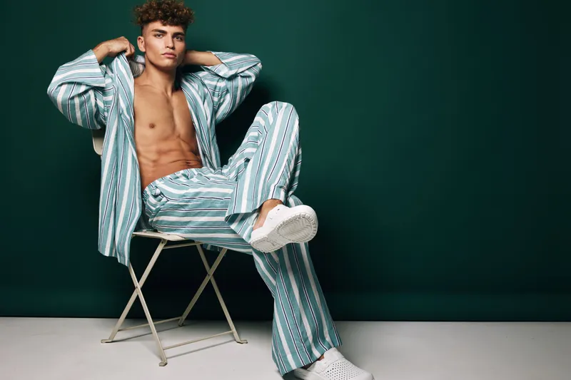 Young man sitting on chair with striped pants and open shirt showing bare chest in studio with dark green background. Casual fashion pose with confident expression and relaxed posture.