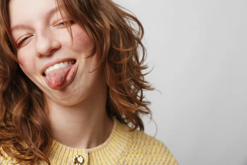 Young woman with curly hair wearing yellow sweater sticks out tongue playfully. Close-up portrait on light background. Concept of fun, youth, carefree attitude, and casual style.