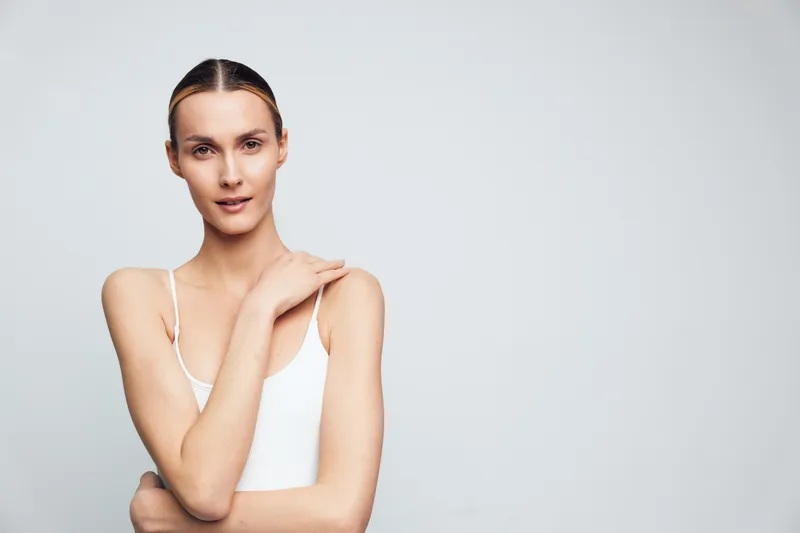 A young woman with clear skin and pulled-back hair poses confidently in a white tank top against a plain light background, conveying natural beauty and simplicity.
