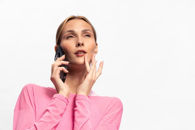 Young woman in pink sweater talks on smartphone with thoughtful expression, isolated on white background. Concept of communication, technology, and casual lifestyle.
