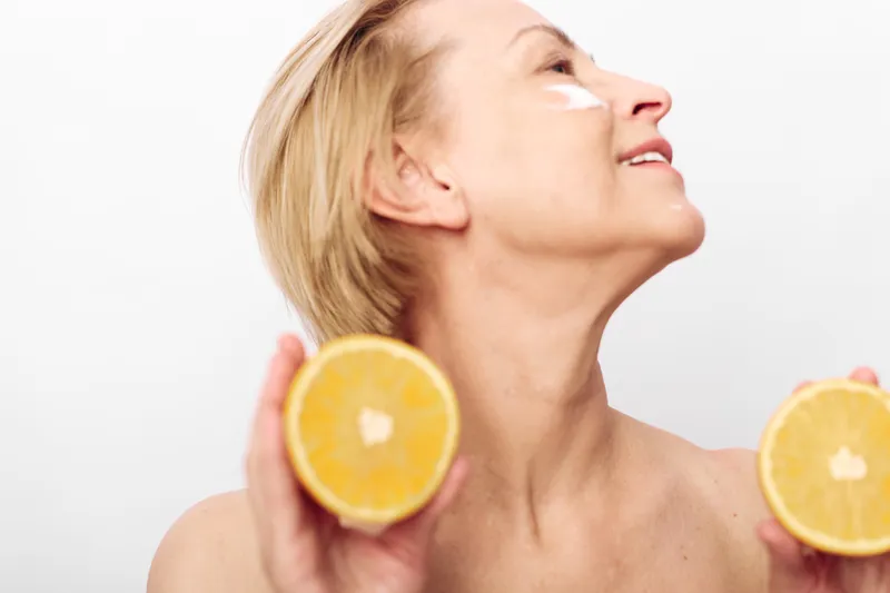 Mature woman with short blonde hair holds two halves of fresh orange in hands, applying white cream on face. Concept of natural skincare, vitamin c, healthy lifestyle and beauty care.