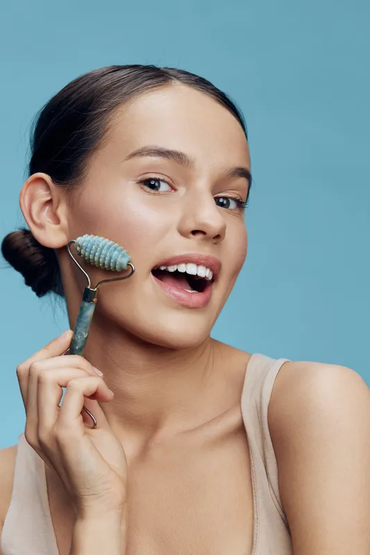 Young woman demonstrating facial roller technique on her skin, showcasing beauty skincare trends and healthy skincare routine against a soft blue background
