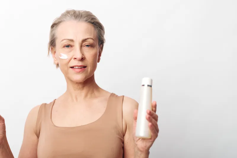Mature woman with short grey hair applying skincare cream under eyes holds white bottle. Beauty concept for anti aging, facial care, wellness, and healthy skin routine on white background.