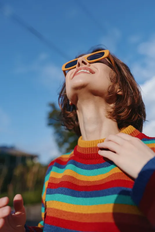 Woman smiling in striped sweater and orange sunglasses, short hair and looking up at sunny sky outdoors. Young adult enjoying travel lifestyle, joyful relaxed pose in warm light.
