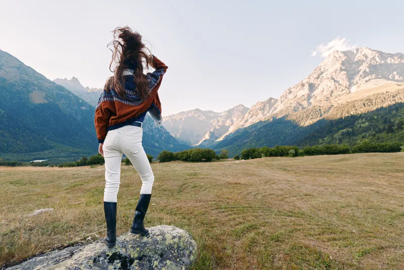 Woman stands on a rock in a wide field looking at jagged mountains, landscape vista in backview wearing a warm sweater and boots, enjoying outdoor solitude and scenic alpine valley.