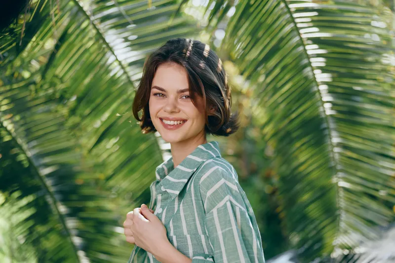 Smiling woman with short dark hair wearing a striped green shirt stands in a sunny tropical setting among palm fronds.