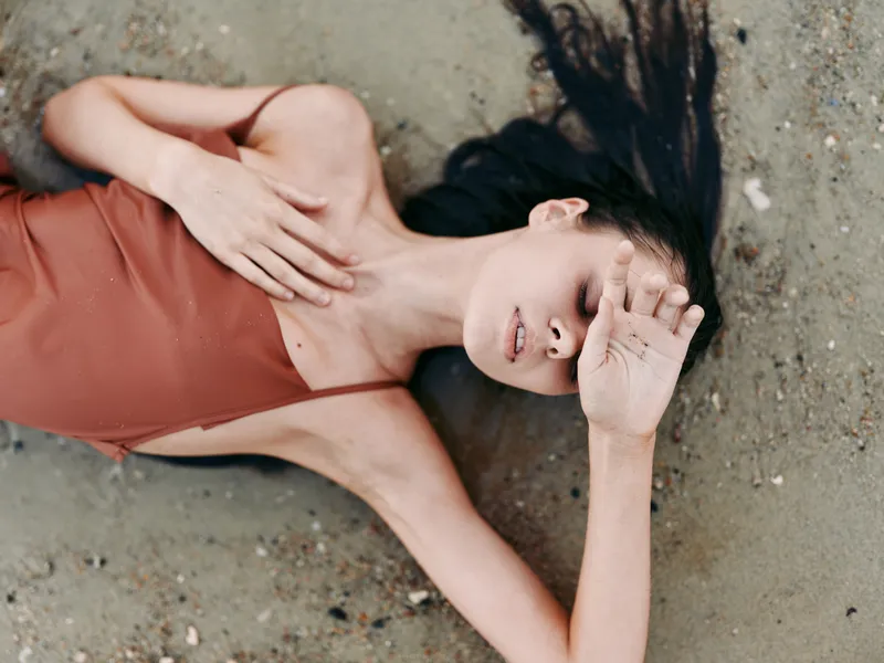 A woman lies on a sandy beach in a rust dress, hair spread across the sand, calm expression, and soft light highlighting curves and a sense of quiet vulnerability.