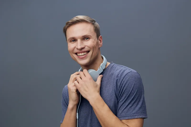 Smiling man, young adult, casual clothing, headphones around neck, friendly expression, studio portrait, gray background, happiness, positive emotions, lifestyle image, male model, cheerful mood