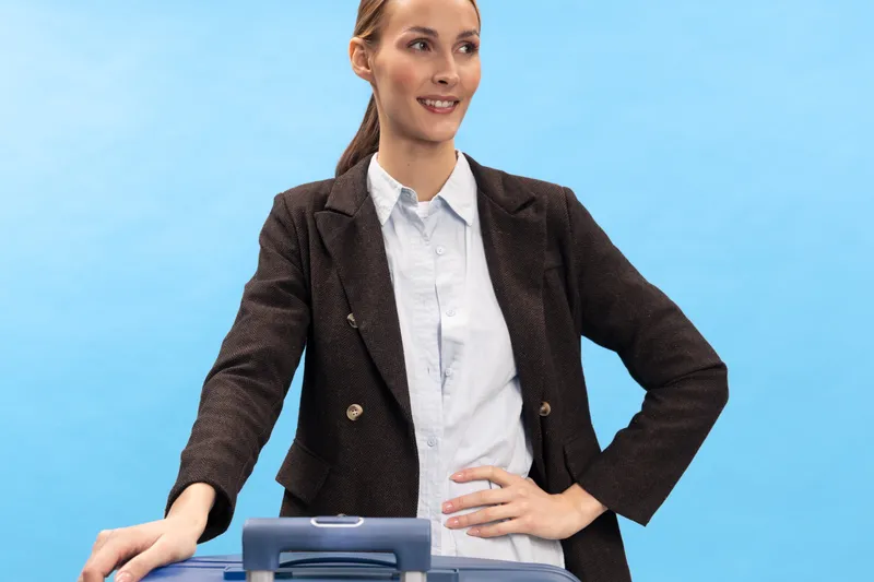 Confident businesswoman in dark blazer stands with hand on suitcase handle, smiling against blue background. Concept of travel, business trip, professionalism, and female traveler.