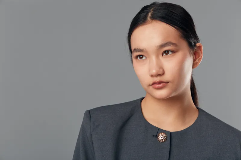 Young woman with dark hair in professional suit looking away thoughtfully on gray background. Portrait of businesswoman in minimal makeup with neat hairstyle and serious expression.
